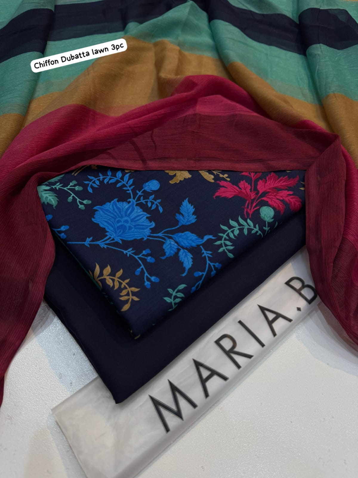 Stack of colorful fabrics with a floral-patterned fabric on top, branded 'Maria B'.