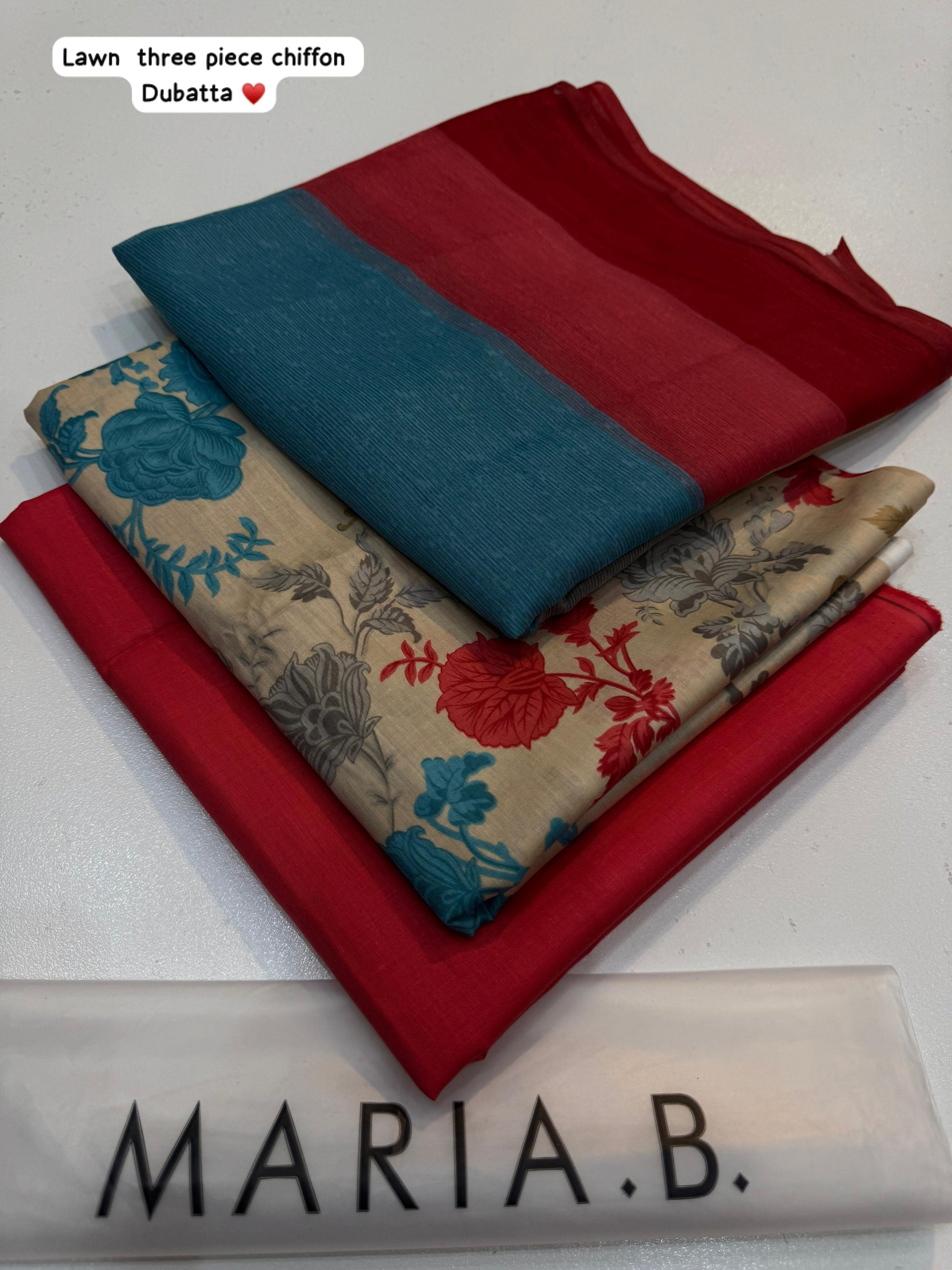 Three folded fabrics in red, blue, and floral pattern on a white surface with 'Maria B.' branding.